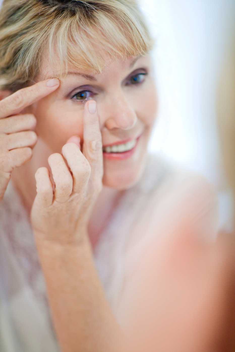 Senior woman looking at her reflection in the mirror while putting in contact lenses.