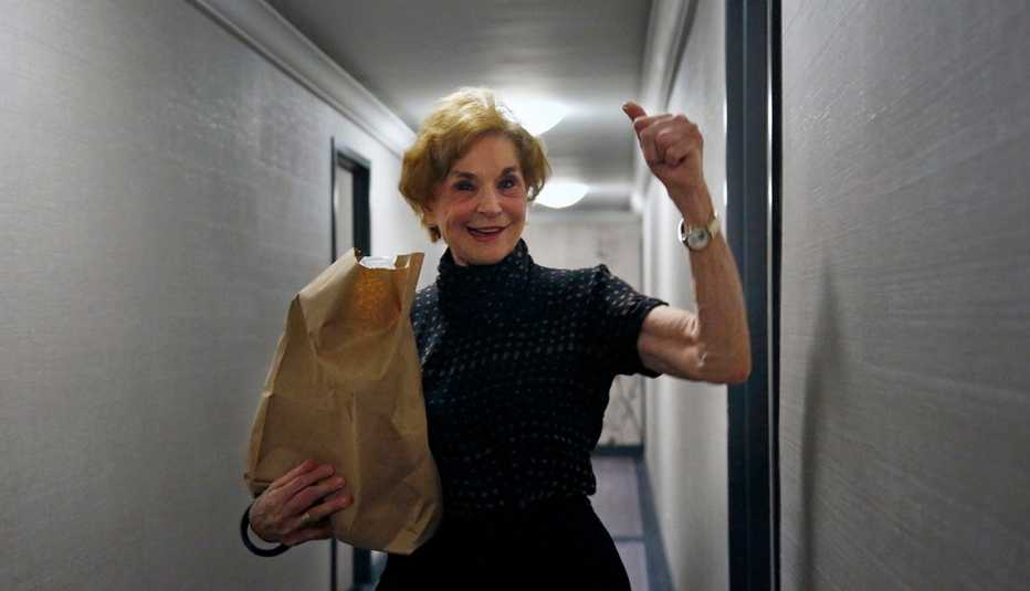 In this March 17, 2020, photo, Carol Sterling, 83, gives a thumbs-up to Liam Elkind after he delivers groceries to her apartment as part of a newly formed volunteer group he cofounded, Invisible Hands. The retired arts administrator has been sheltering at