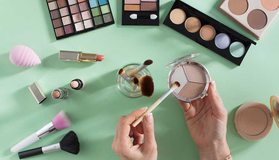 Various types of makeup on a green background. Woman's hands holding eyeshadow and a brush.
