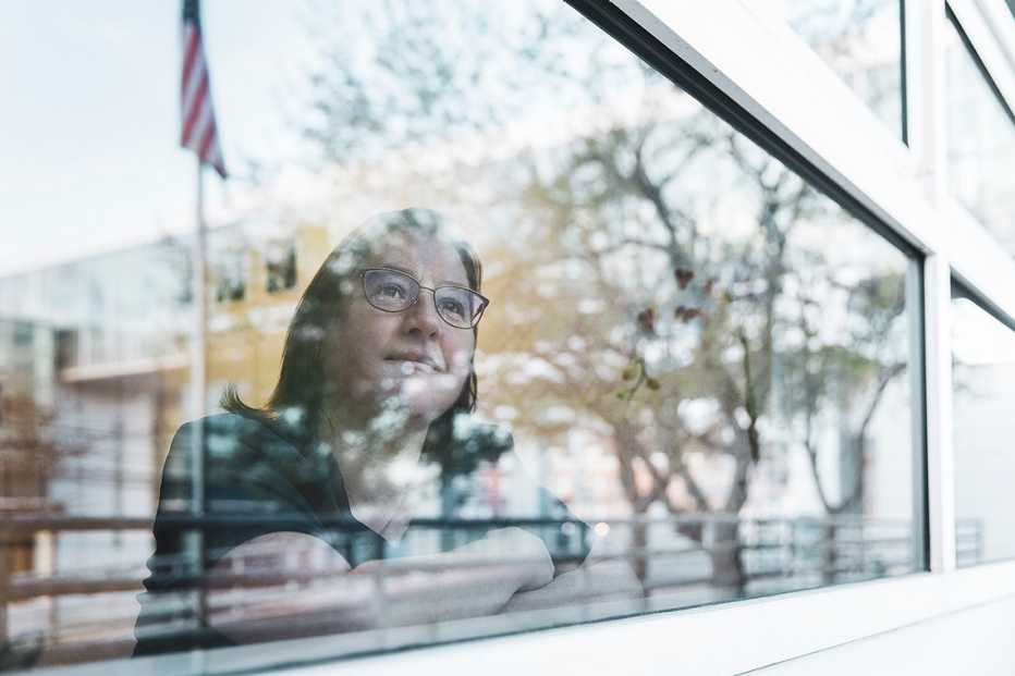 a nurse looks at a window shot form outside partially obscured by the trees and an american flag on a pole reflected in the window 
