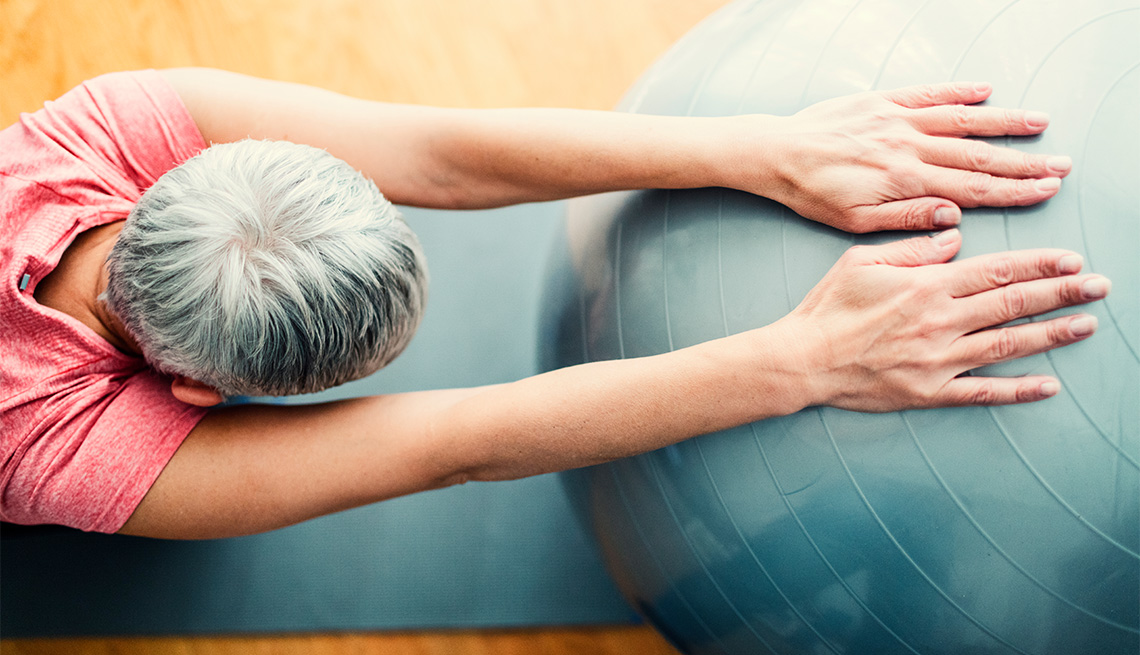 Mature woman exercising at home. Sitting on exercise mat and working stretching exercise while holding hands on pilates ball. Overhead view.