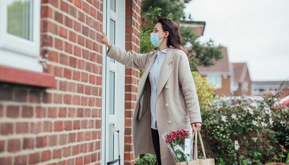 A woman wearing a face mask knocking on the front door of a home holding a bag and flowers