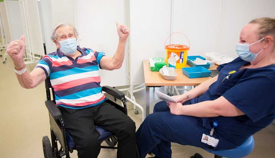 man in a wheel chair raising his hands in joy after receiving a coronavirus vaccine. a healthcare working sitting next to him is laughing