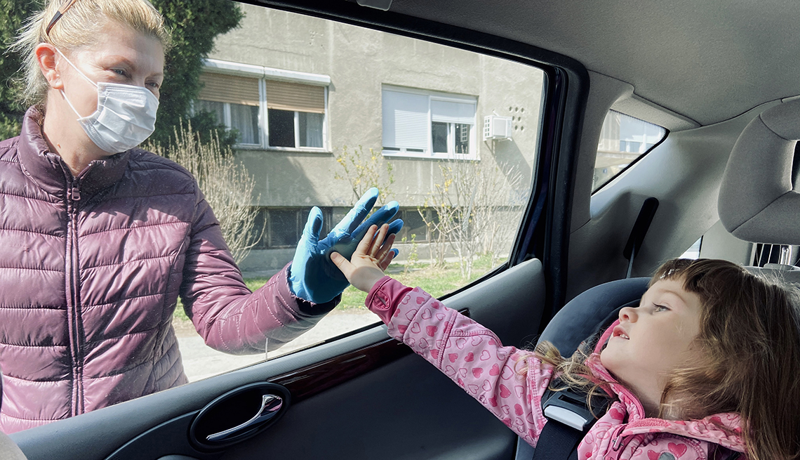 Woman touching her grandchild's hand through a car window, wearing a face mask