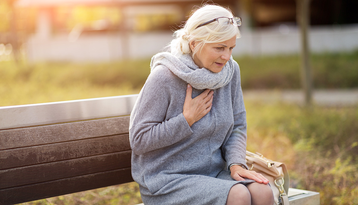 Older woman wearing a gray scarf and dress sitting on a bench holding her chest due to tightness and shortness of breath