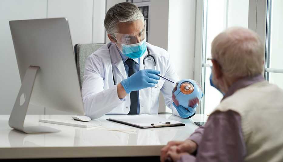 Doctor pointing to a model of an eye while a male patient watches. Both are wearing face masks.