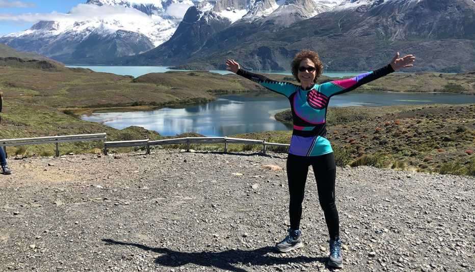 a smiling woman stands on gravel with her arms raised up in front of a mountain range and a lake