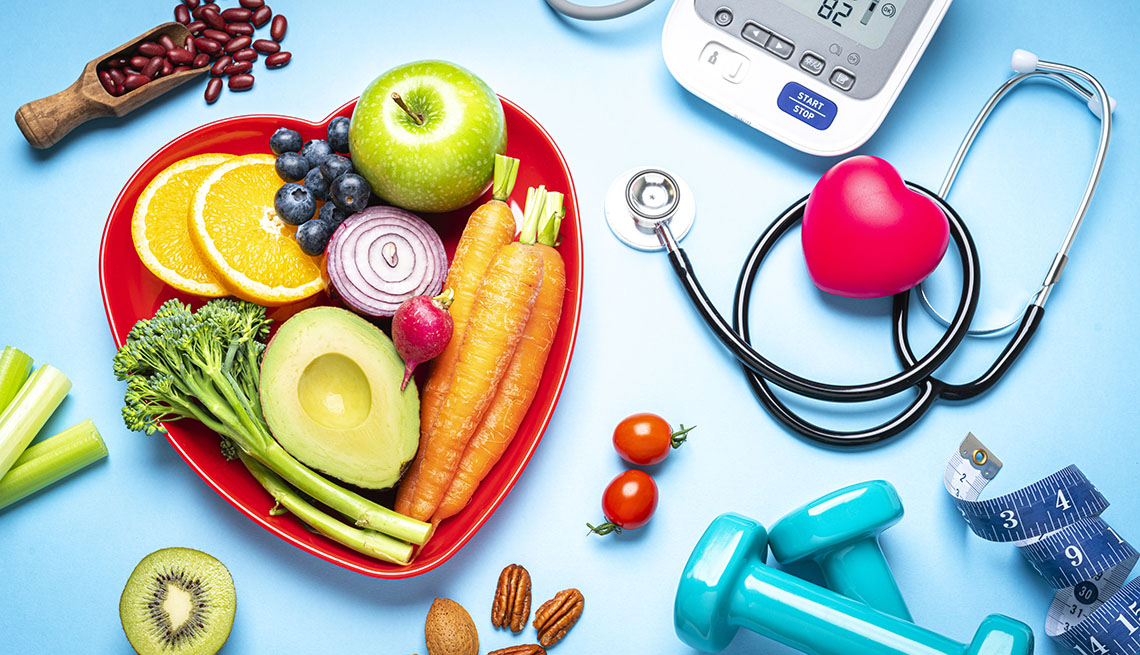 red heart shape plate with fresh organic fruits and vegetables shot on blue background. A digital blood pressure monitor, doctor stethoscope, dumbbells and tape measure are beside the plate 