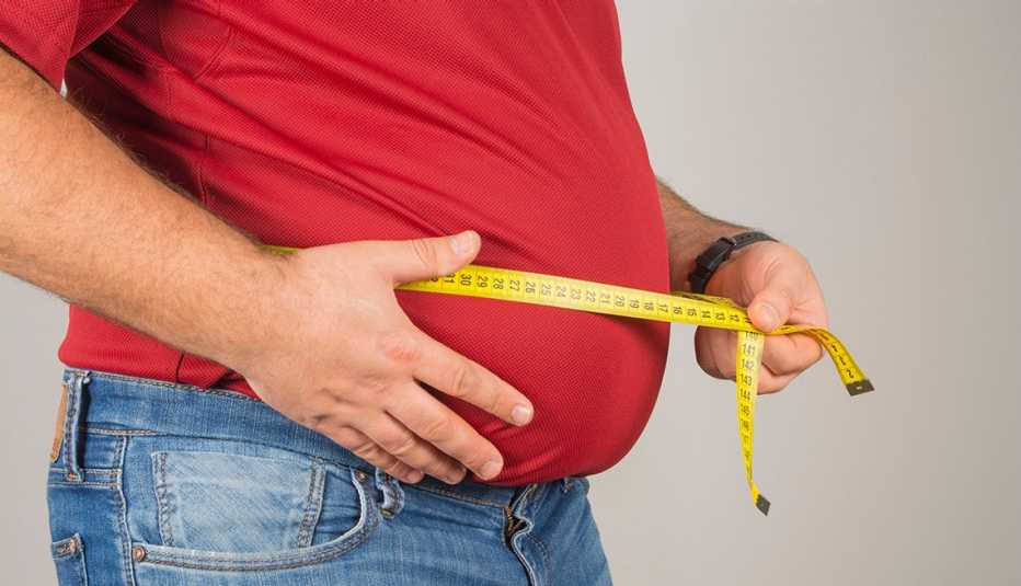 a man in a red t-shirt and jeans measures his belly with a yellow tape measure