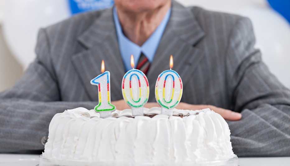 A birthday cake with lit candles for a one hundredth birthday. Older man in suit sits behind the cake.