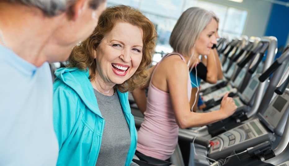 Mature woman working out in a gym, talking to a man who is also working out.  Another woman working out in the background.
