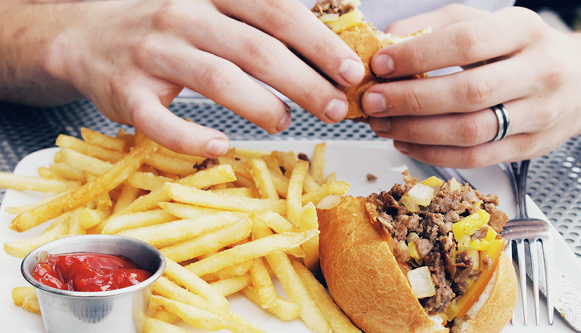 hands holding cheese steak sandwich above plate with fries