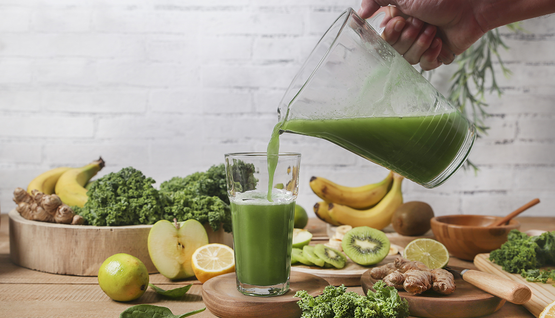 A person pouring green smoothie into a glass which is surrounded by fruits and vegetables