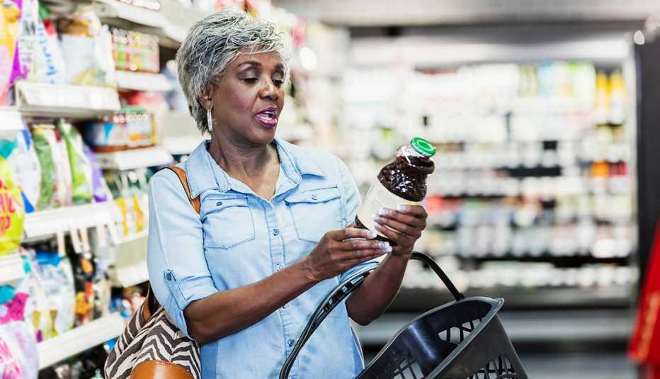 A senior African-American woman in her 60s shopping in a grocery store, carrying a shopping basket. She is reading the nutrition label on a bottle.