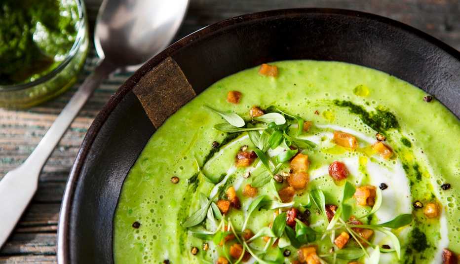 A bowl of avocado soup with bacon and mint pesto on a table with a metal spoon nearby