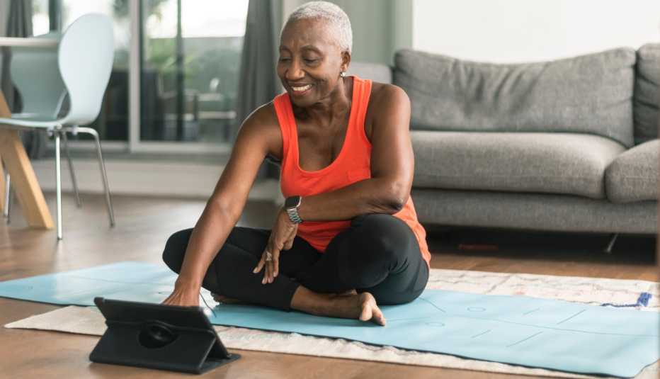 a smiling woman sits on a blue yoga mat and uses her tablet to do an online yoga class at home