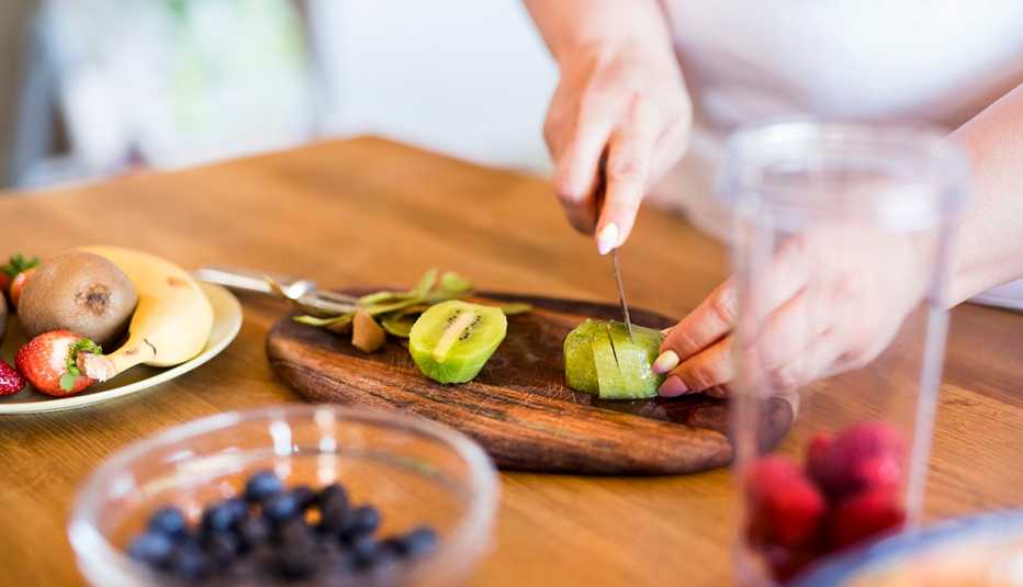 woman cutting kiwi on a cutting board with other foods, including blueberries, in bowls nearby