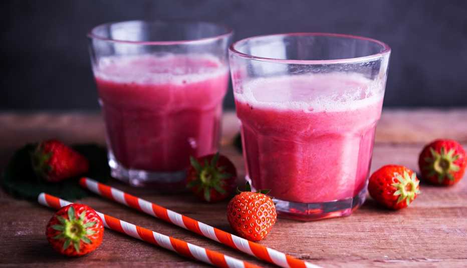 close up of two strawberry smoothies for healthy weight gain surrounded by red and white striped straws and fresh whole strawberries on a kitchen table