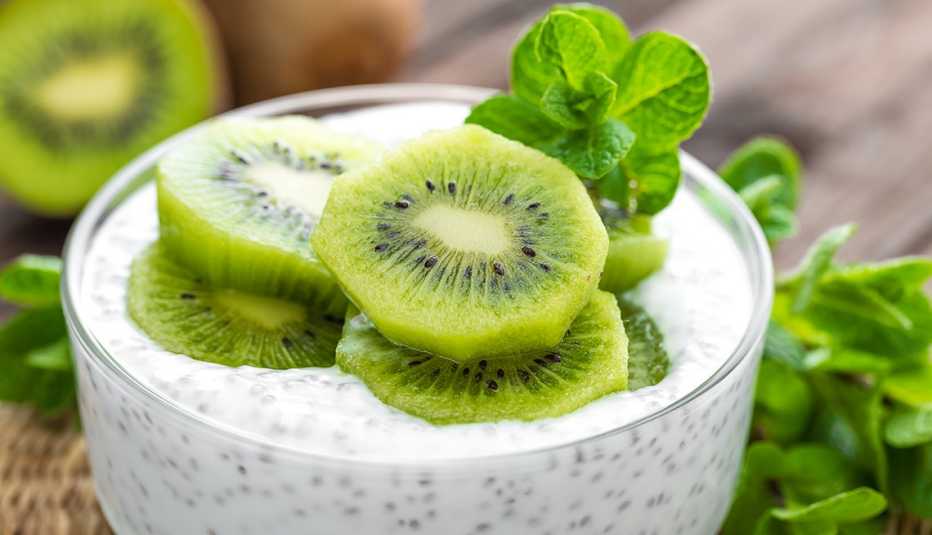 close up of a high-fiber bowl of chia pudding topped with slices of kiwi and a sprig of mint on a kitchen table with a fiber-rich kiwi in the background