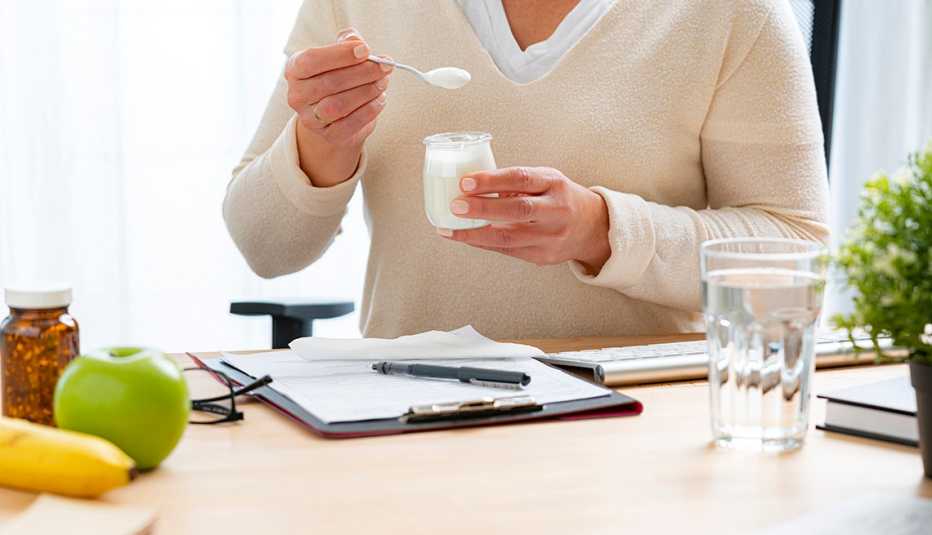 woman at her desk eating yogurt and drinking water, fruit and vitamins are nearby.