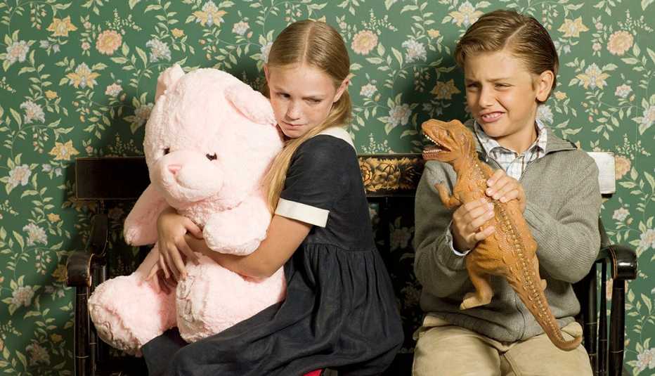 Young Female Sibling Holds Large Pink Teddy Bear And Frowns As Her Brother Teases Her With Toy Dinosaur On Bench, Indoors, AARP Home And Family, Family And Friends, Can Bullying Between Brothers and Sisters Ever Stop?  