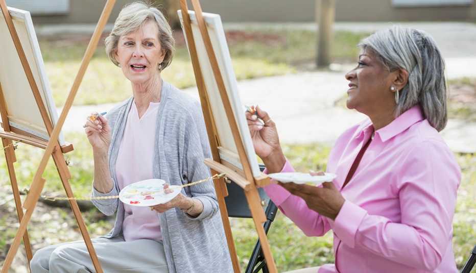 Two women taking an art class outdoors. They are sitting at easels, painting on a canvas. 