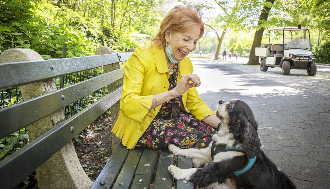 woman sitting on park bench holding a treat for her dog who has its front paws up on the bench looking at her