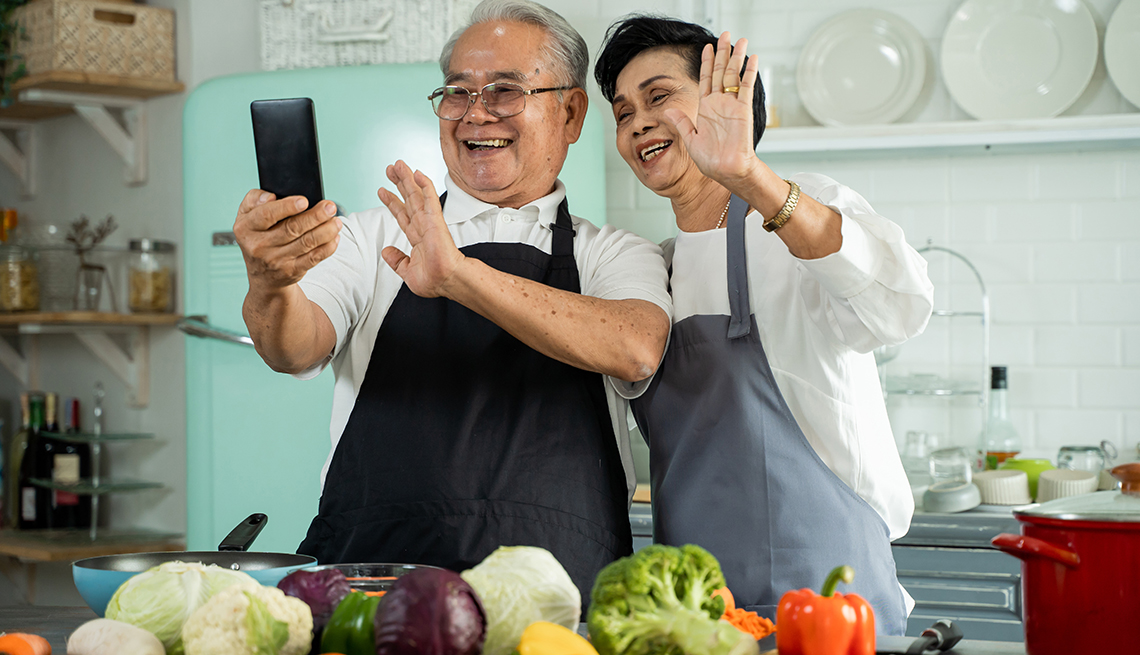 A man and woman stand in a kitchen waving at a phone