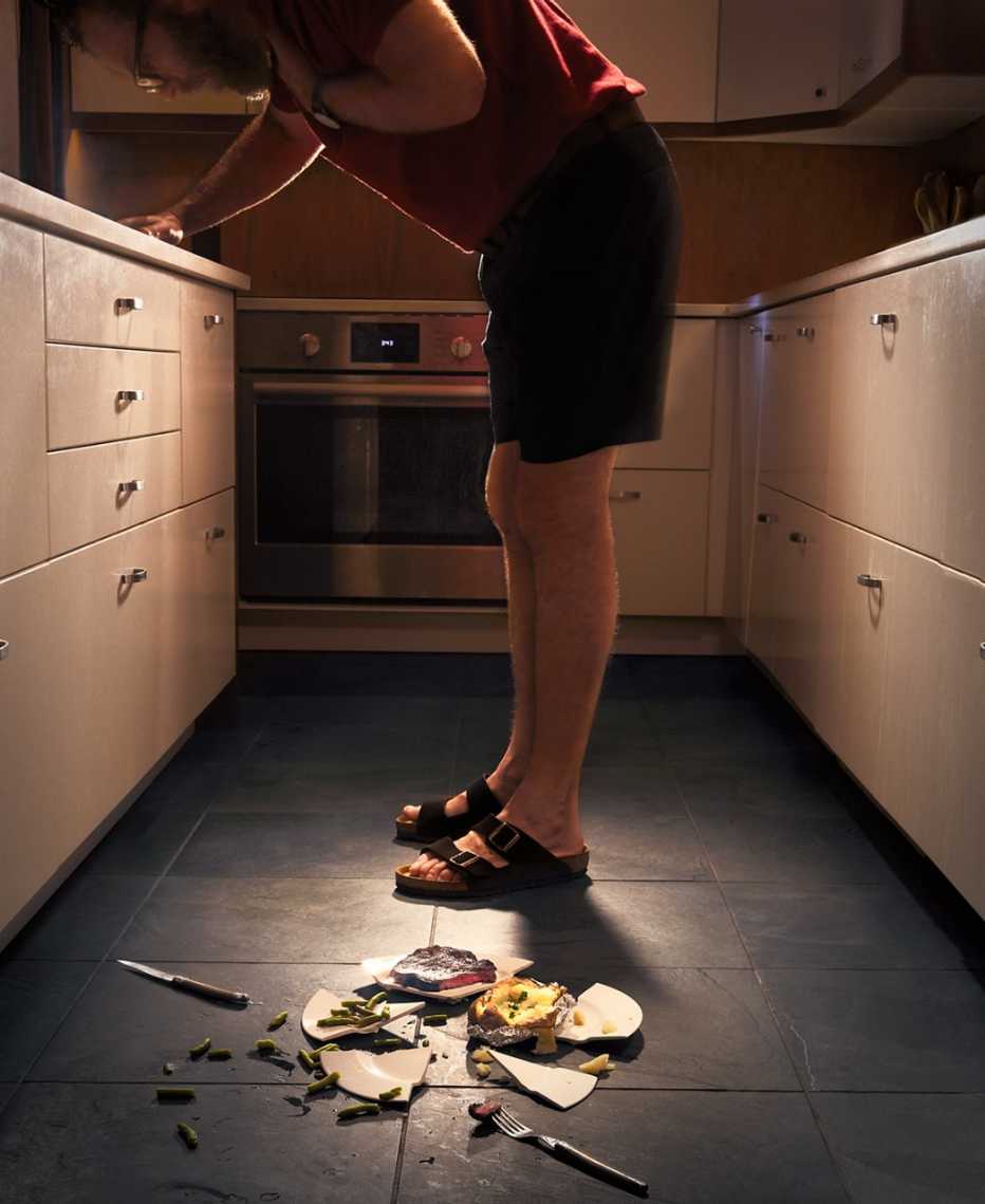 man in his kitchen bending over and choking on food, there is a broken plate of food at his feet