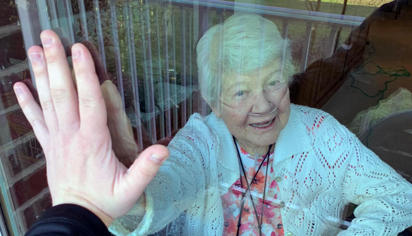a nursing home resident and a person outside the window touch hands on either side of the glass