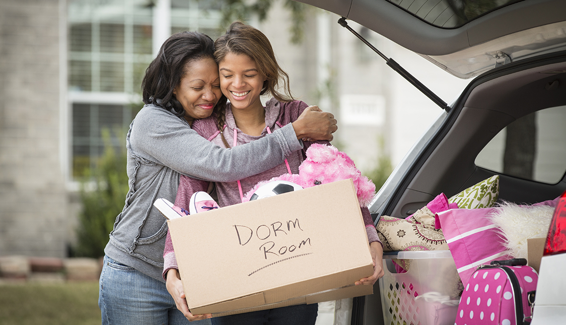 A young adult loading a box that says dorm room on it into her car as her mother hugs her