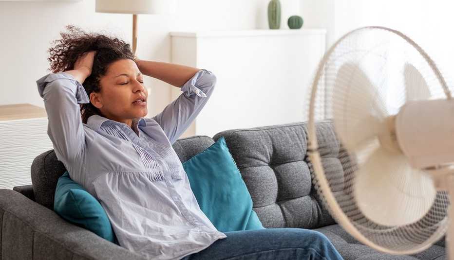 Woman sits on sofa holding hair off her face to feel the cooling breeze from an electric fan on the coffee table