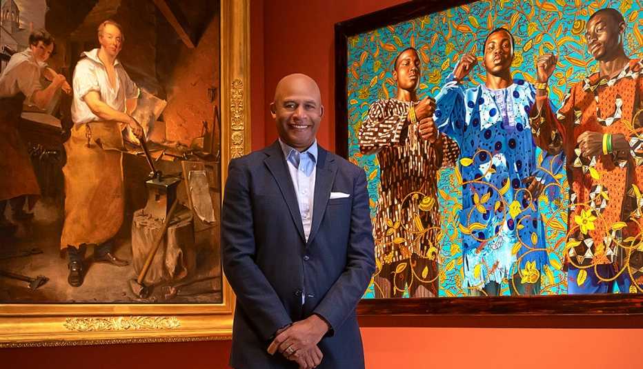 eric pryor the president of the pennsylvania academy of fine arts tands in front of two paintings on the left is pat lyon at the forge by john neagle and on the right is three wise men greeting entry into lagos by kehinda wiley