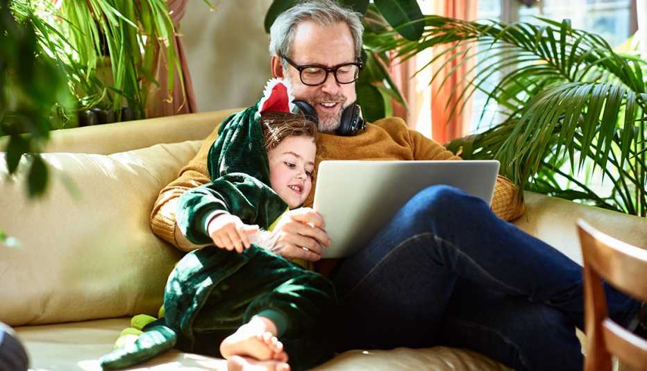 a grandfather sits on a couch looking at laptop with his grandchild