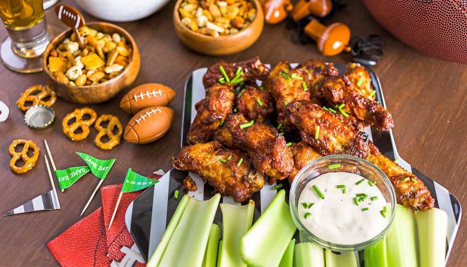 A Superbowl party table with chicken wings, celery, and dip, party pretzel snack mix, banners, beer, and a football.