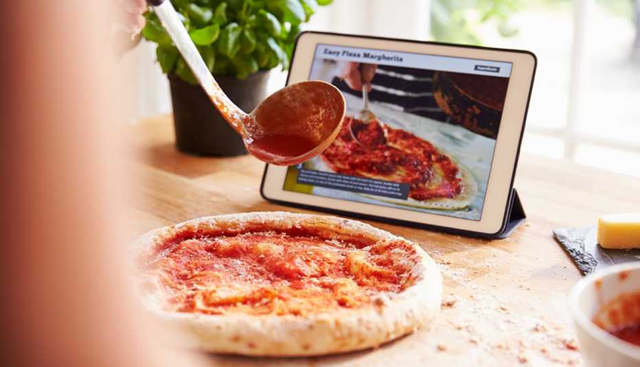 a person partially off-camera pours sauce on pizza dough while following a recipe on a tablet