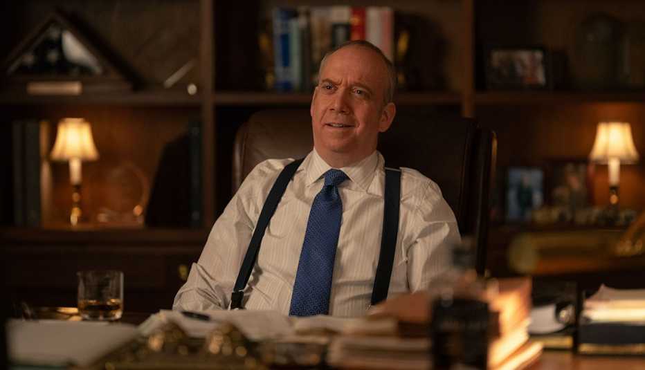 billions star paul giamatti wears a tie and suspenders over a white button up shirt while sitting at a desk on the set of billions