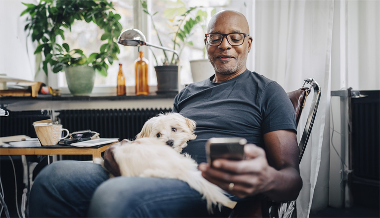 a man sitting at his dinning room table looking at a mobile phone as his dog sits on his lap