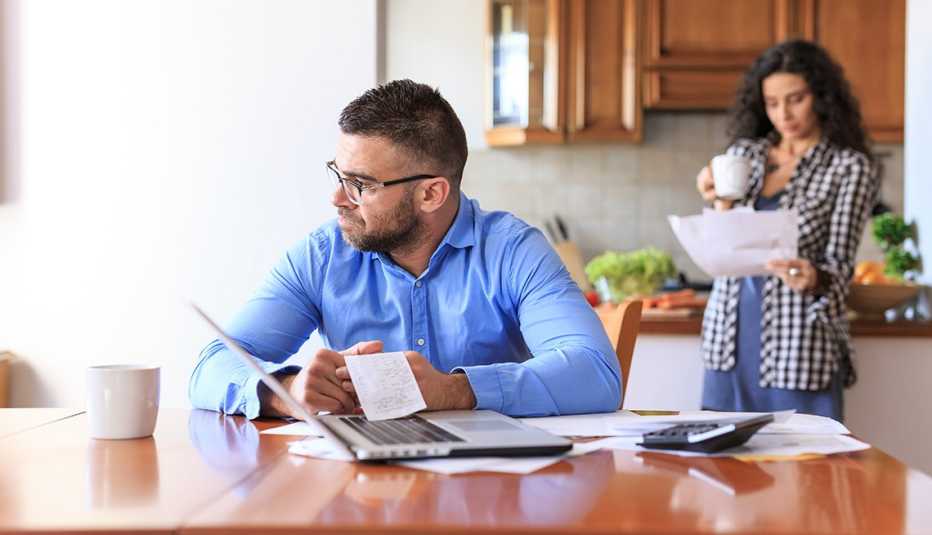 Man sitting at a table reviewing bills and receipts with woman in the background reviewing papers