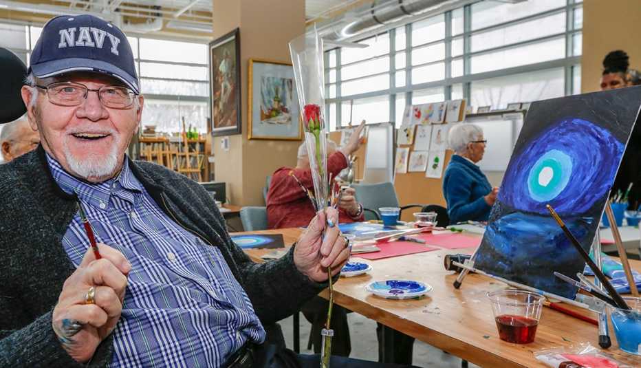 older man wearing a navy baseball hat holds a rose while sitting in an art class in front of a table top easel