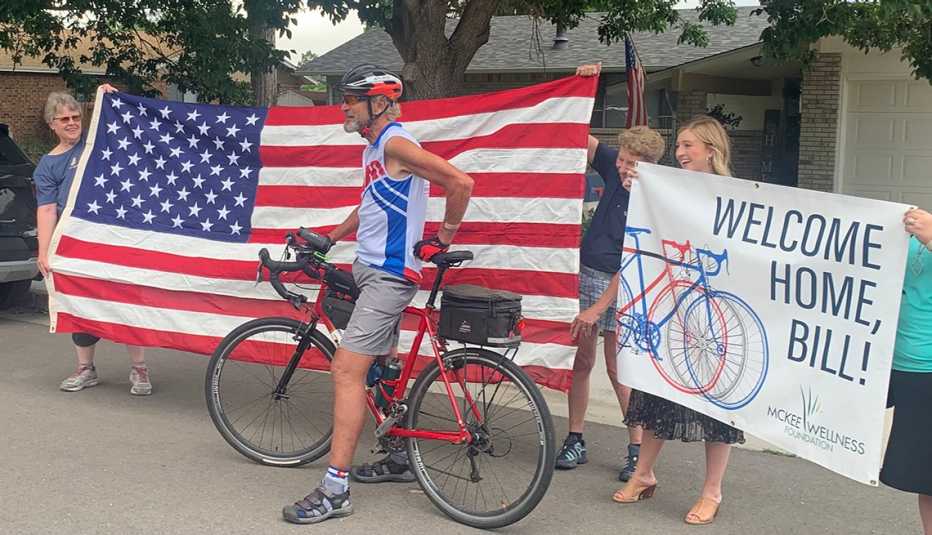 bill wallace sits on a red bicycle while friends and family hold an american flag and a welcome home, bill sign behind him