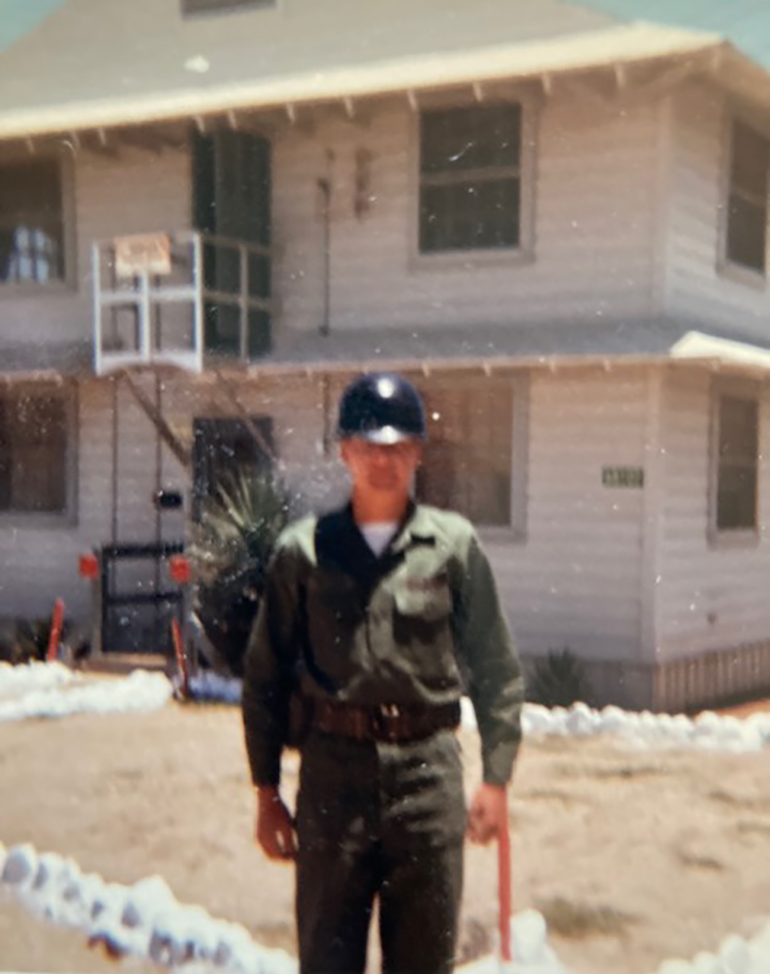a young bill wallace stands in an army uniform and helmet in front of a white building