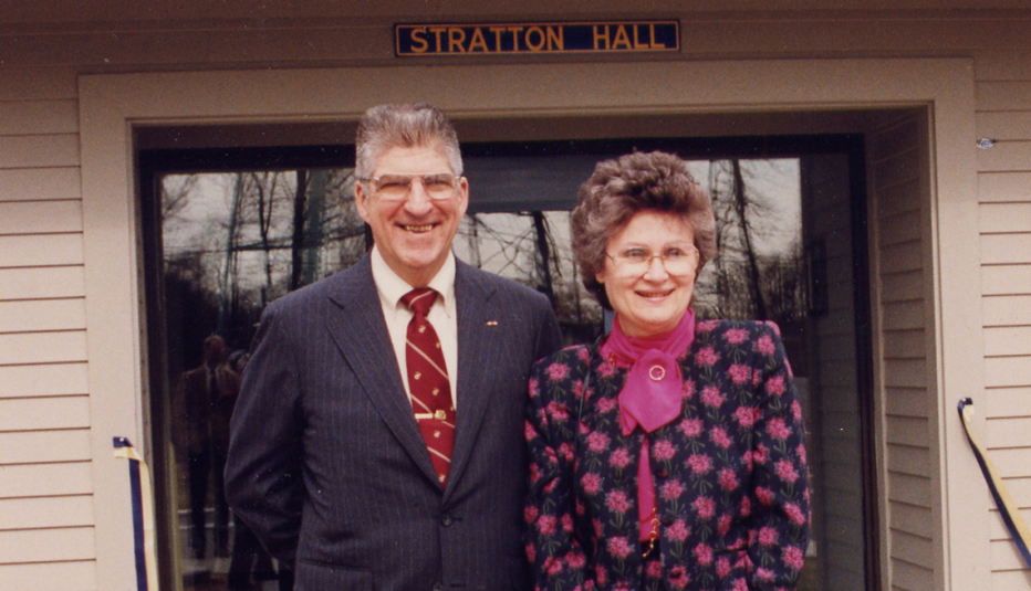 Dick and Alice Stratton stand in front of a set of doors, with a sign saying Stratton Hall above them.