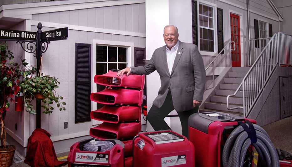 a smiling man in a light shirt, gray blazer and dark pants stands with red PuroClean machines outside a home