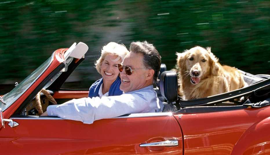 an older couple smiles while driving in a red convertible with a golden retriever in the backseat