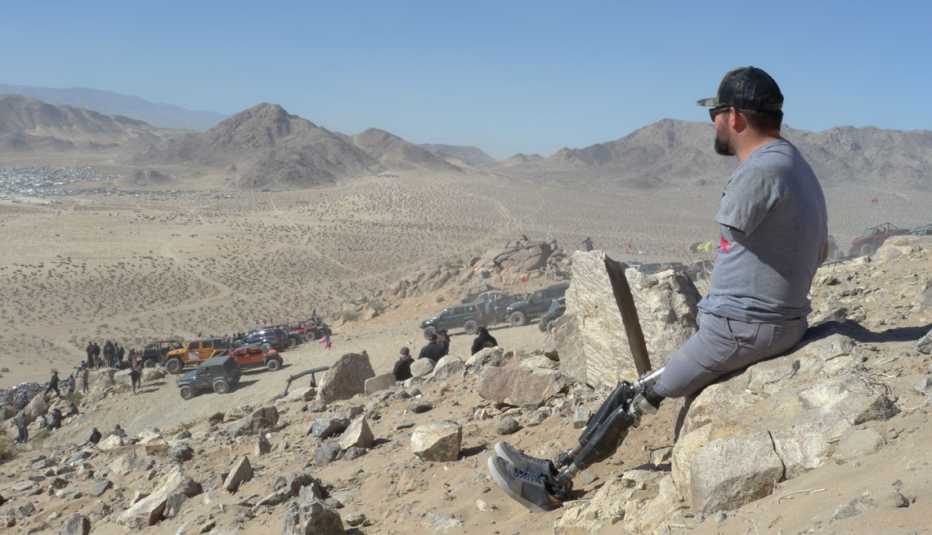 a man sits atop a rock formation, looking out at the desert. he is a triple amputee after losing both legs and his left arm