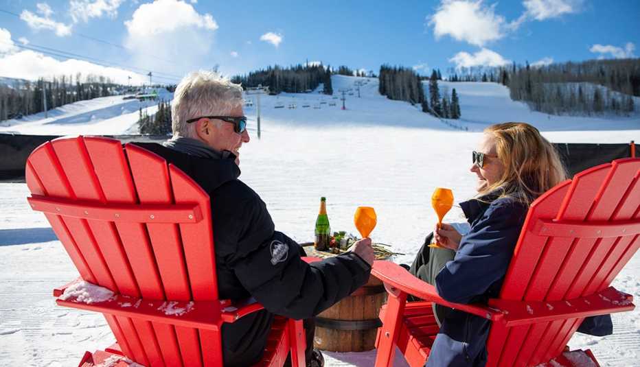 a smiling man and woman sit in red chairs and enjoy drinks while sitting beneath a slope at a ski resort
