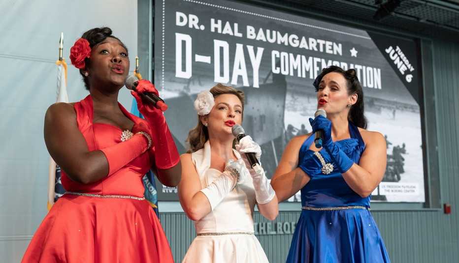 three women in nineteen forties style red, white, and blue dresses sing in front of a d day commemoration sign