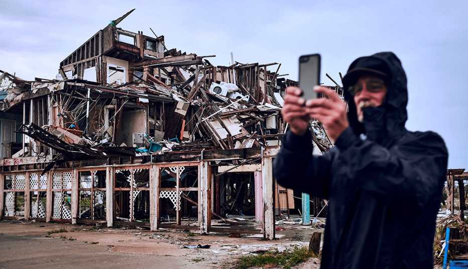 A man in a black hooded coat holds up his smartphone in front of a building destroyed by a storm