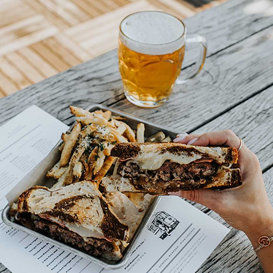 close up of a patty melt sandwich with french fries and a beer on a table